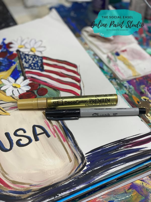 A gold paint pen and a black sharpie marker on a mixed media pad. On the Mixed media pad there is a mason jar with USA painted on the front. Inside of the jar are colorful flowers and an american flag. 